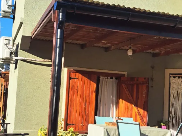 Detalle de una galería con techo de madera, canaleta oscura y poste, frente a una ventana con postigos.