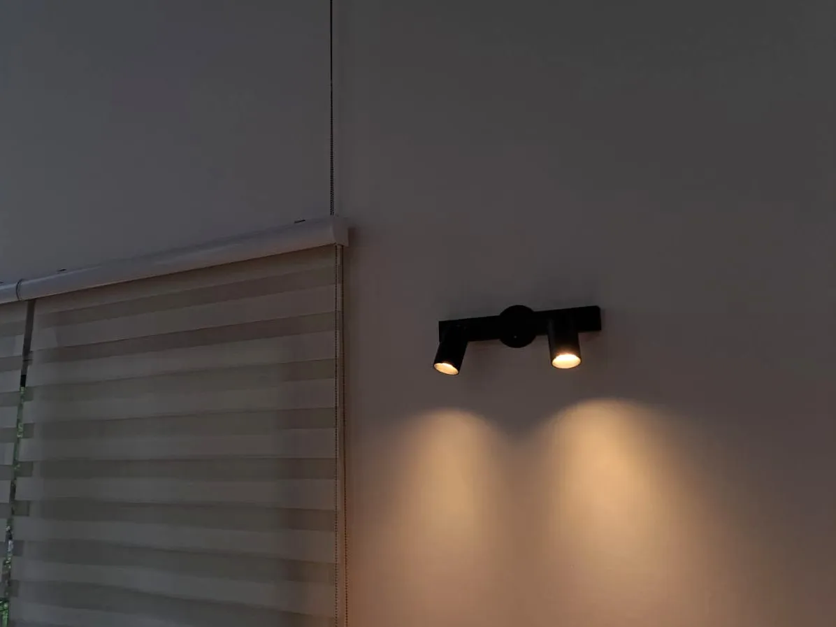 Aplique negro de dos focos encendidos sobre una pared clara, junto a una ventana cubierta por una cortina.