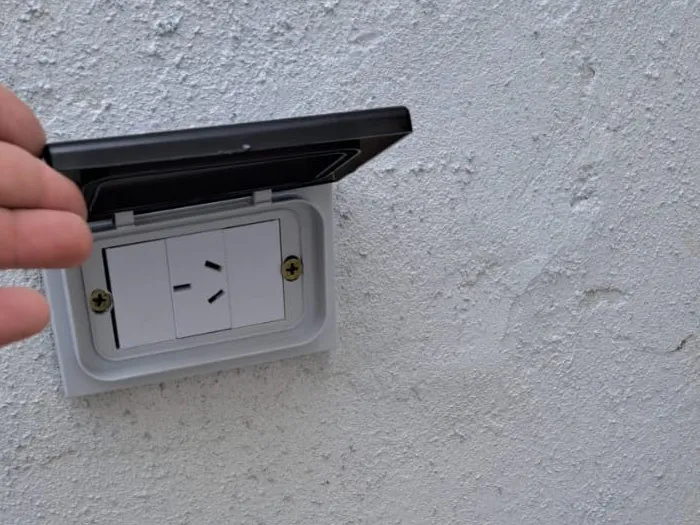 Tomacorriente exterior con tapa de protección levantada y dedos de una mano visibles sobre una pared gris.