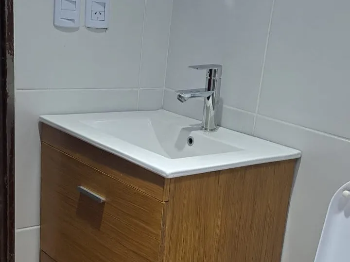 Bacha blanca con grifería monocomando cromada sobre un vanitory de madera, junto a una pared revestida.